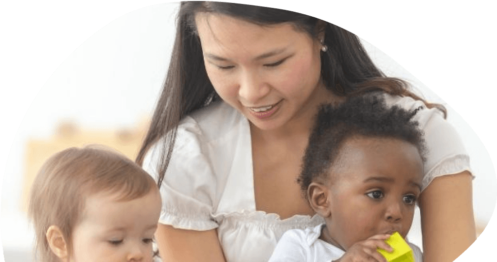 A caregiver in a yellow sweater leaning over a toddler girl in a purple top as they play with wooden blocks on a table.