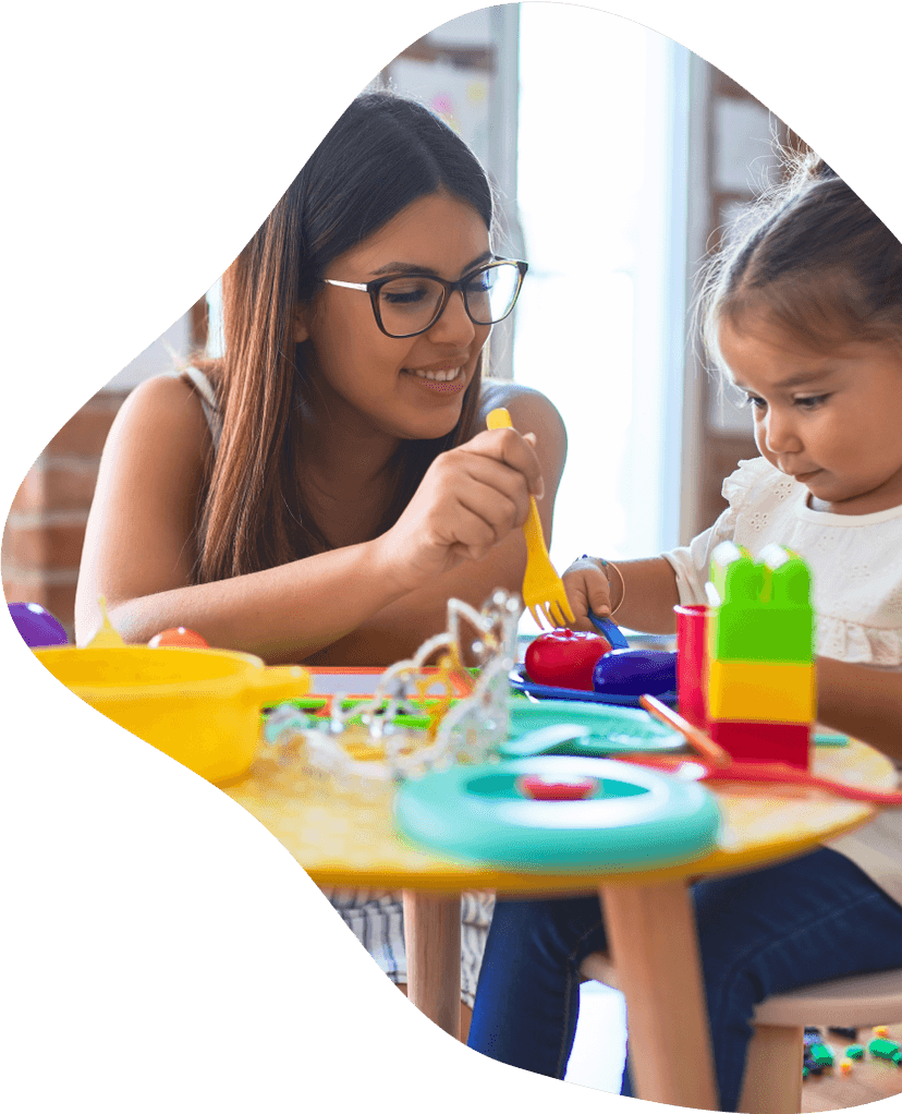 A caregiver in a yellow sweater leaning over a toddler girl in a purple top as they play with wooden blocks on a table.