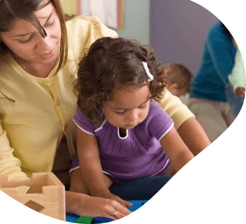 A caregiver in a yellow sweater leaning over a toddler girl in a purple top as they play with wooden blocks on a table.